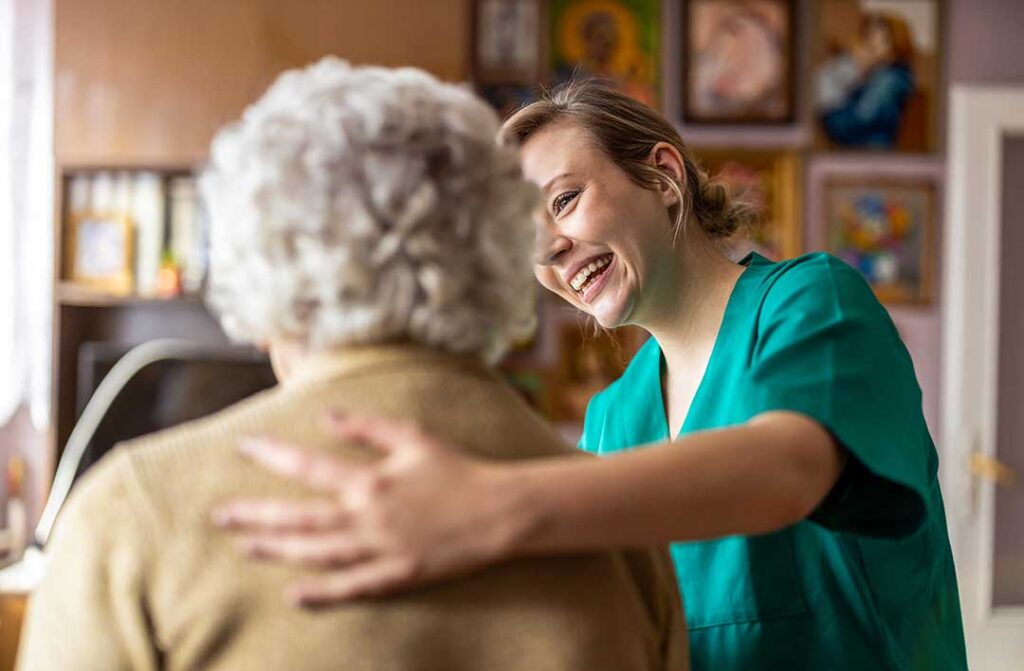 Caregiver assisting an older adult in a bright assisted living hallway while another staff member checks a care chart.