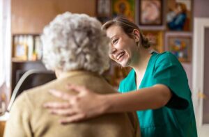 Caregiver assisting an older adult in a bright assisted living hallway while another staff member checks a care chart.